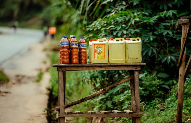 Rural roadside stall featuring bottles and jerrycans of oil and juice with a lush green backdrop.