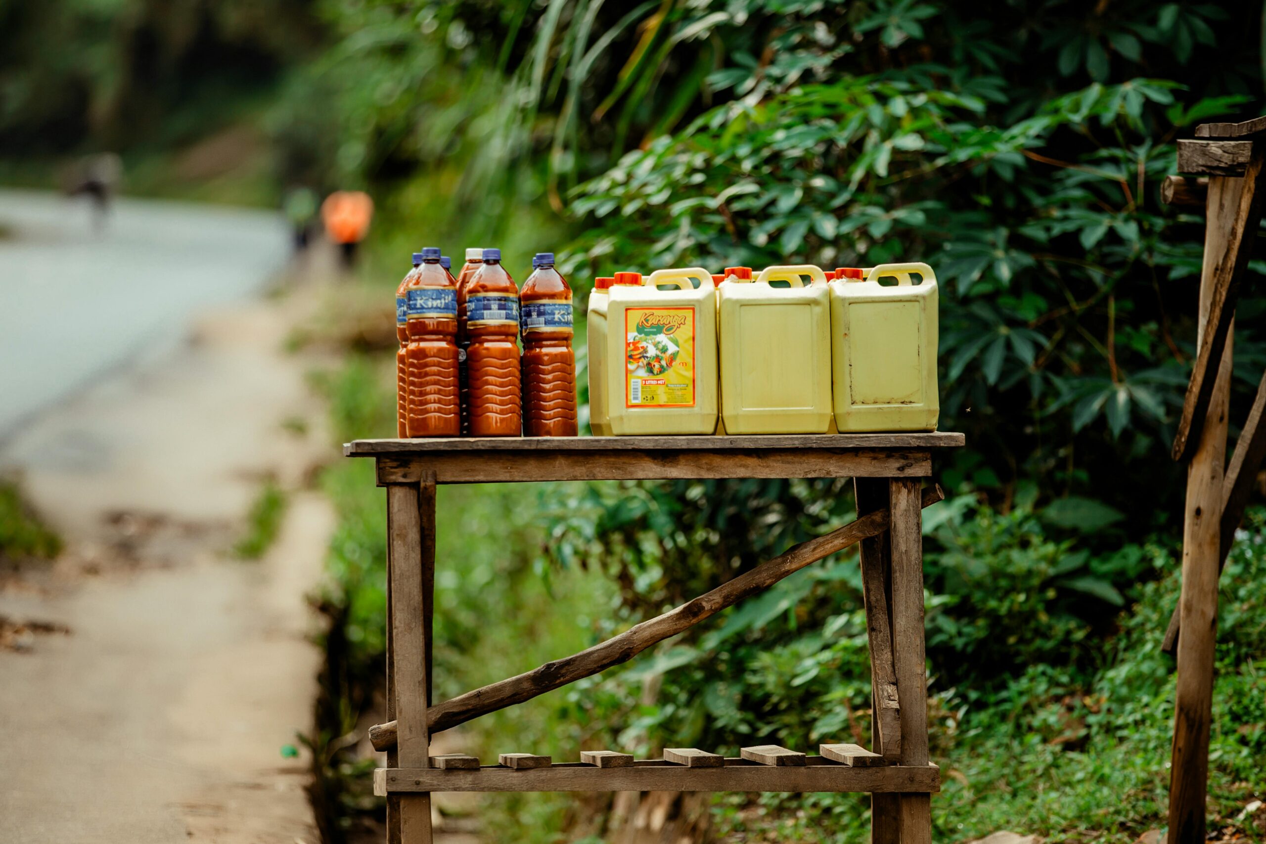 Rural roadside stall featuring bottles and jerrycans of oil and juice with a lush green backdrop.